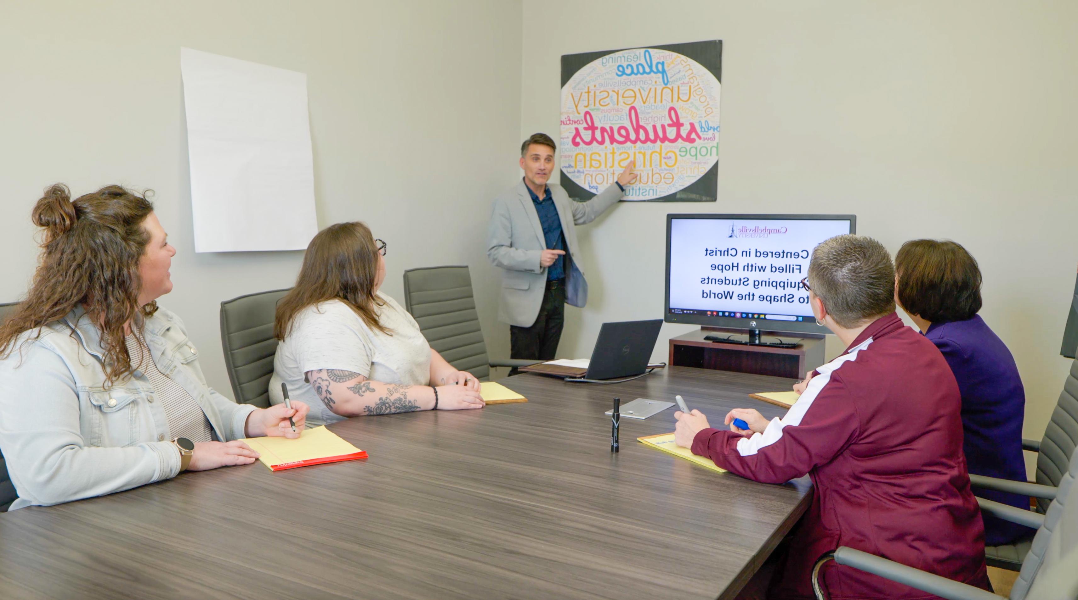 A group of people gathered around a table listening to a presenter.